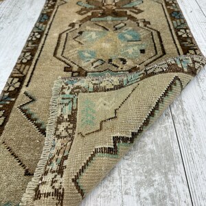 May include: A rectangular, vintage rug in beige and brown tones. It features a central design with floral and geometric patterns in brown and teal. The edges are bordered with a darker brown pattern, and the rug has a fringed edge. The rug is folded over.