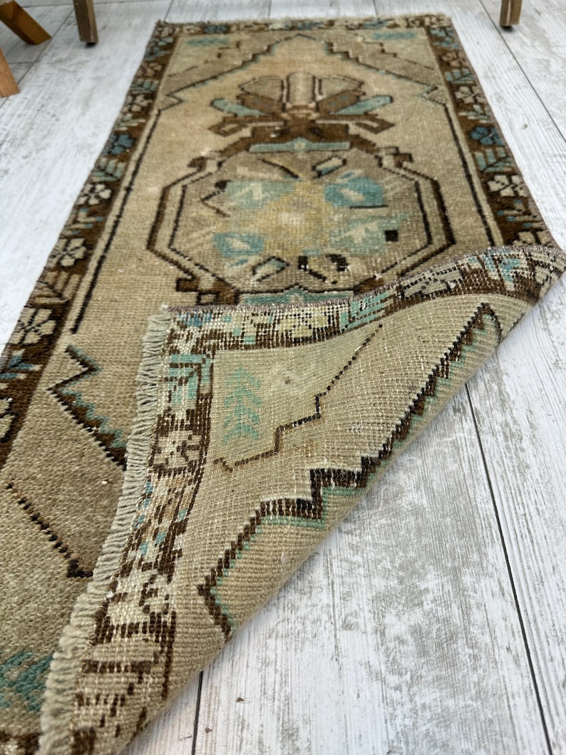 May include: A rectangular, vintage rug in beige and brown tones. It features a central design with floral and geometric patterns in brown and teal. The edges are bordered with a darker brown pattern, and the rug has a fringed edge. The rug is folded over.
