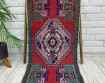May include: A rectangular woven rug featuring a vibrant red background with a large central diamond motif in light purple. The design includes geometric patterns in blue, green, yellow, and dark brown, finished with a dark border along the edges.