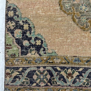 May include: A woven rug featuring a tan background with a dark blue and teal floral border. The central area displays a large, intricate medallion pattern in shades of dark blue, teal, and mustard yellow, with a fringed edge at the bottom.