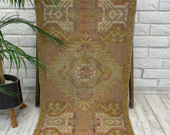 May include: A rectangular woven rug featuring a central diamond medallion pattern in muted shades of tan, olive green, and faded pink. The rug has a textured pile with geometric motifs and a fringed edge, displaying a weathered, vintage appearance.