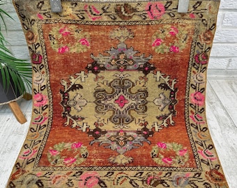 May include: A square rug featuring a central beige medallion with dark brown and pink geometric patterns, surrounded by a deep orange field. The border displays a repeating floral motif in pink, green, and brown, finished with a fringed top edge.