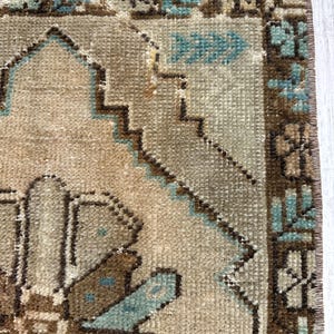 May include: Close-up of a beige and brown rug with a geometric pattern. The rug features a central design with brown and blue accents, and a border with similar colours. The rug has a distressed, vintage look.