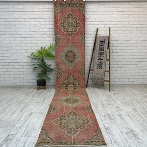 May include: A long, narrow runner rug featuring a faded red base with repeating intricate floral medallion patterns in muted green, brown, and cream tones. The rug is displayed vertically, with a matching smaller piece draped over a wooden ladder.