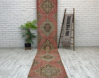 May include: A long, narrow runner rug featuring a faded red base with repeating intricate floral medallion patterns in muted green, brown, and cream tones. The rug is displayed vertically, with a matching smaller piece draped over a wooden ladder.