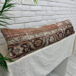 May include: A long, rectangular pillow with a vintage rug design. The pillow features a mix of rust, beige, and brown tones with intricate patterns. It rests on a white surface against a white brick wall, showcasing a rustic aesthetic.