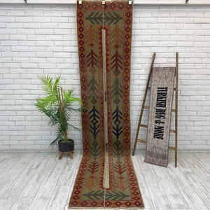 May include: A long, narrow runner rug featuring a muted olive green background with a repeating pattern of stylized tree motifs in orange, dark blue, and green. The rug has a dark brown border with a geometric diamond pattern throughout the design.