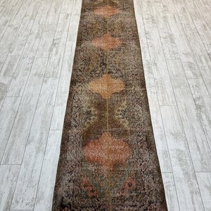 May include: A long, narrow runner rug featuring a distressed, weathered appearance with a central vertical seam. The design displays repeating orange, quatrefoil-shaped medallions set against a dark, muted background of brown, grey, and faded green tones.