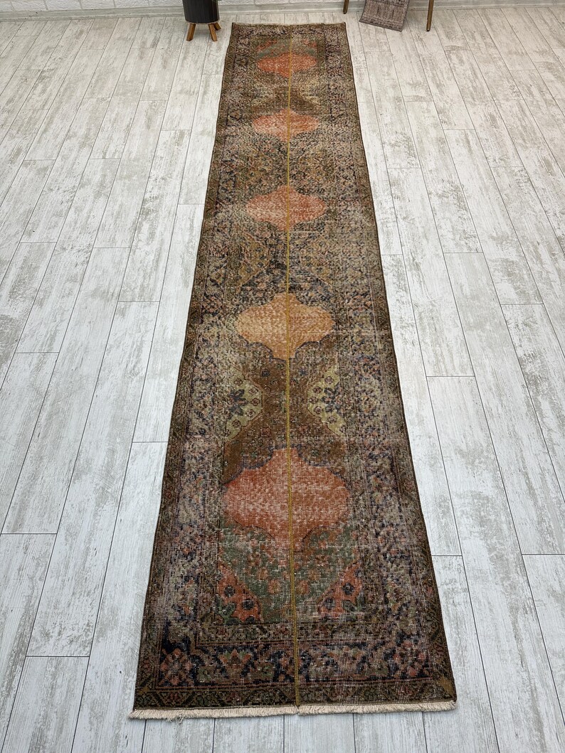 May include: A long, narrow runner rug featuring a distressed, weathered appearance with a central vertical seam. The design displays repeating orange, quatrefoil-shaped medallions set against a dark, muted background of brown, grey, and faded green tones.