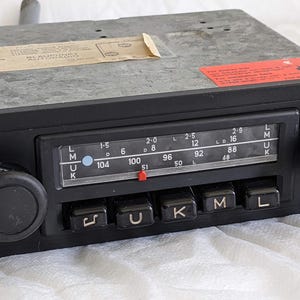 Puede incluir: Una radio de coche rectangular negra con una pantalla analógica horizontal de frecuencias con texto blanco, dos perillas negras circulares a los lados y cinco botones negros con símbolos y las letras U, K, M y L en el panel frontal.