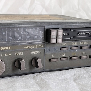 Puede incluir: Un autorradio con reproductor de casetes Blaupunkt Nashville R24 en color gris oscuro, con pantalla analógica de sintonización FM y AM, ranura para casetes con la etiqueta DNR-SYSTEM, mandos de volumen, graves y agudos, y botones de función.