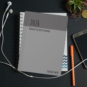 Könnte beinhalten: Ein graues Notizbuch mit dem Text "2026 Burnout Recovery Journal" und dem Namen "Georgia Smith". Das Notizbuch liegt auf einem Spiralblock. Kopfhörer, ein Smartphone und ein Stift sind ebenfalls zu sehen.