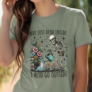 Pode incluir: Uma camiseta verde com um esqueleto regando um vaso de flores. O texto na camiseta diz "Not just dead inside I also go outside".