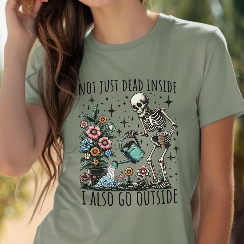 Pode incluir: Uma camiseta verde com um esqueleto regando um vaso de flores. O texto na camiseta diz "Not just dead inside I also go outside".