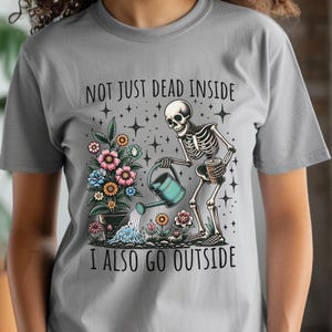 Pode incluir: Uma camiseta cinza com um esqueleto regando um vaso de flores. O texto na camiseta diz "Not just dead inside I also go outside".