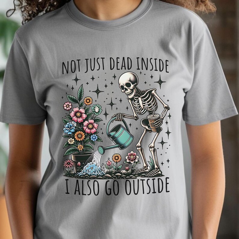 Pode incluir: Uma camiseta cinza com um esqueleto regando um vaso de flores. O texto na camiseta diz "Not just dead inside I also go outside".