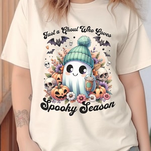 Peut inclure: T-shirt crème avec un fantôme de dessin animé portant un bonnet vert et tenant un café. Le motif comprend des citrouilles, des fleurs, des crânes et des chauves-souris, avec le texte "Just a Ghoul Who Loves Spooky Season."