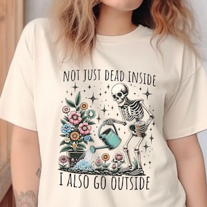 Pode incluir: Uma camiseta bege com um gr&aacute;fico de um esqueleto regando um vaso de flores. O texto "Not just dead inside I also go outside" est&aacute; impresso na camiseta.