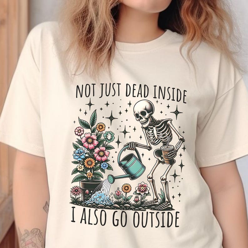 Pode incluir: Uma camiseta bege com um gr&aacute;fico de um esqueleto regando um vaso de flores. O texto "Not just dead inside I also go outside" est&aacute; impresso na camiseta.