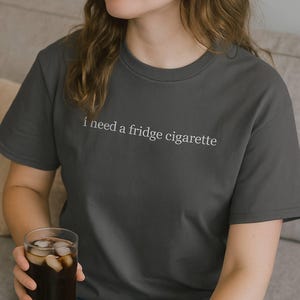 Puede incluir: Camiseta gris oscuro con la frase blanca "i need a fridge cigarette". La camiseta es de cuello redondo y está hecha de un material suave. Se sostiene un vaso de bebida helada en la mano.