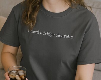 Pode incluir: T-shirt cinza escuro com a frase branca "i need a fridge cigarette". A camiseta é de gola redonda e é feita de um material macio. Um copo de bebida gelada é segurado na mão.