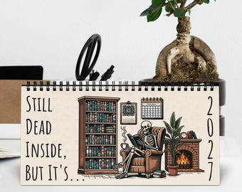 May include: A 2027 spiral-bound desk calendar featuring an illustration of a skeleton sitting in a brown armchair, reading a book, and holding a mug. Next to the skeleton is a tall bookshelf, a fireplace with a fire, and the text Still Dead Inside, But It's.