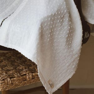 May include: A white blanket featuring a textured swiss dot pattern across the fabric. The blanket has a small rectangular fabric label attached to the bottom corner with visible text and a logo. The material appears lightweight and soft.