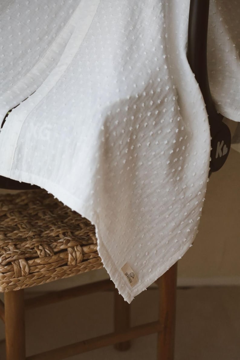 May include: A white blanket featuring a textured swiss dot pattern across the fabric. The blanket has a small rectangular fabric label attached to the bottom corner with visible text and a logo. The material appears lightweight and soft.