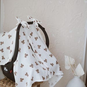 May include: A white muslin car seat cover featuring a repeating pattern of small brown teddy bears. The fabric is draped over a black infant car seat and secured at the top with two tied fabric knots. The material has a soft, textured appearance.