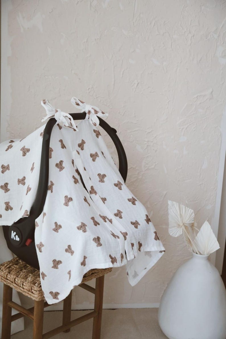 May include: A white muslin car seat cover featuring a repeating pattern of small brown teddy bears. The fabric is draped over a black infant car seat and secured at the top with two tied fabric knots. The material has a soft, textured appearance.