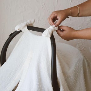 May include: A white, textured swiss dot fabric cover draped over a black infant car seat handle. The fabric features a small, raised dot pattern and is secured to the curved black frame with two tied fabric knots at the top corners.