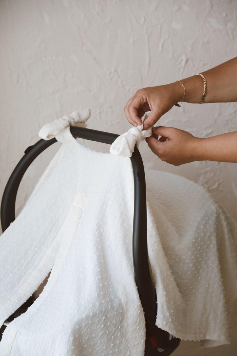 May include: A white, textured swiss dot fabric cover draped over a black infant car seat handle. The fabric features a small, raised dot pattern and is secured to the curved black frame with two tied fabric knots at the top corners.