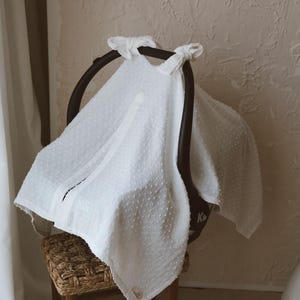 May include: A white textured fabric cover with a subtle dotted pattern draped over a black infant car seat. The cover features two fabric ties at the top handle and a small rectangular label near the bottom edge, providing a soft, layered appearance.