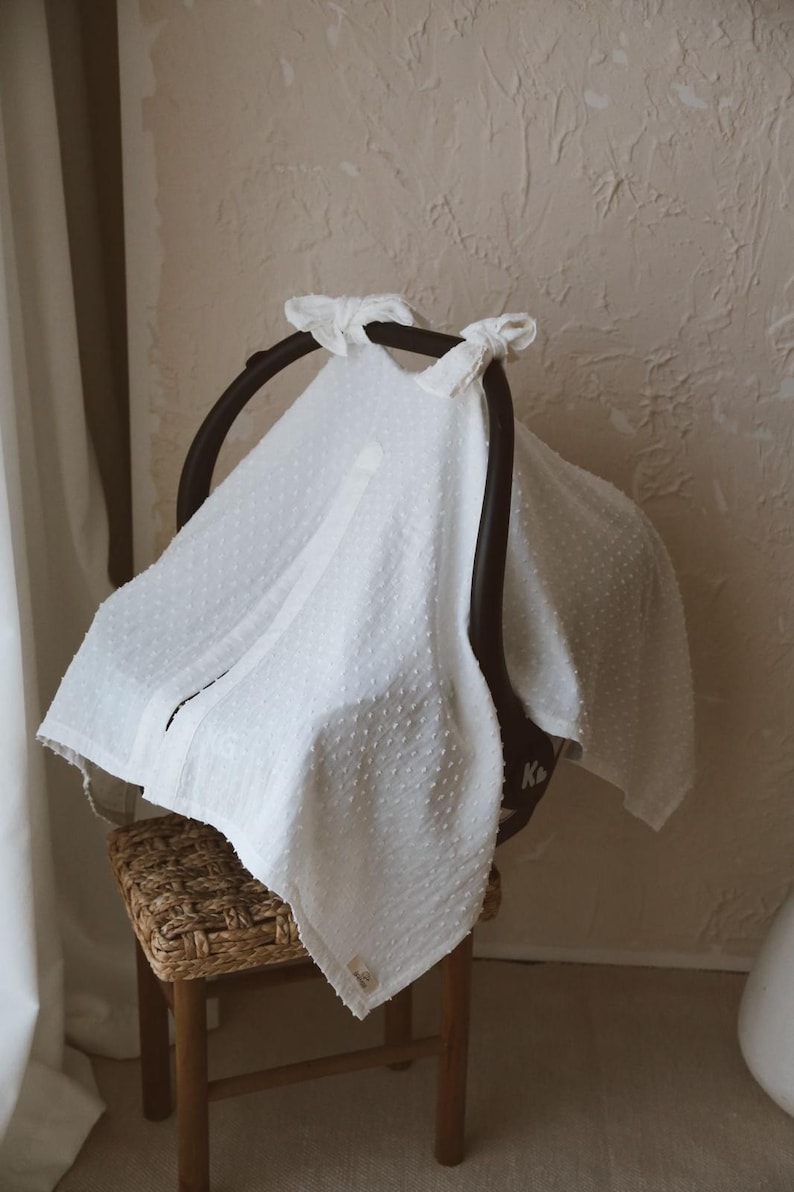 May include: A white textured fabric cover with a subtle dotted pattern draped over a black infant car seat. The cover features two fabric ties at the top handle and a small rectangular label near the bottom edge, providing a soft, layered appearance.