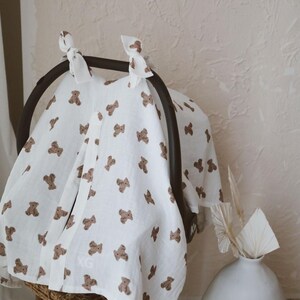 May include: A white muslin car seat cover featuring a repeating pattern of small brown teddy bears. The fabric is draped over a black car seat handle and secured at the top with two white fabric ties. The material has a soft, textured appearance.