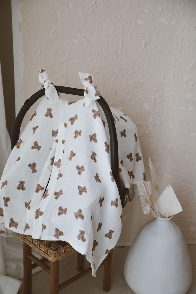 May include: A white muslin car seat cover featuring a repeating pattern of small brown teddy bears. The fabric is draped over a black car seat handle and secured at the top with two white fabric ties. The material has a soft, textured appearance.