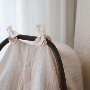 May include: A light-colored, textured fabric cover draped over a black infant car seat handle. The cover features a subtle vertical striped pattern in soft peach and white tones, secured at the top with two tied fabric bows on the handle.