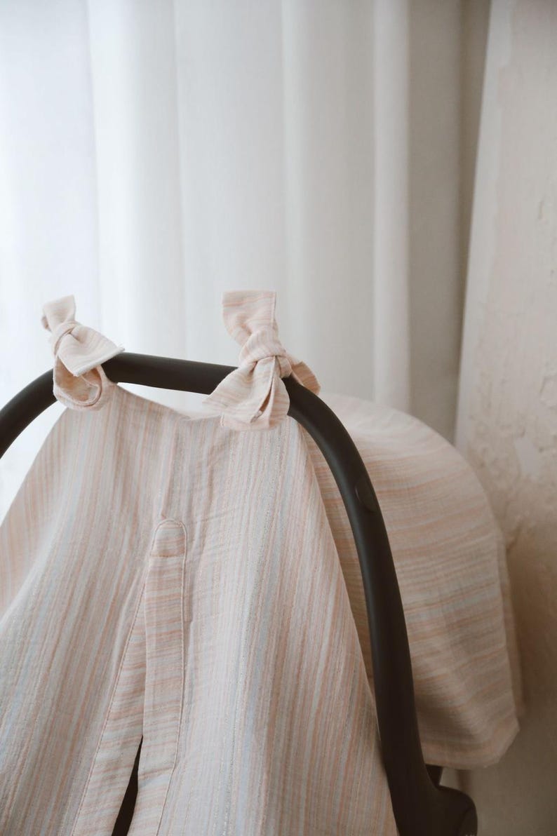 May include: A light-colored, textured fabric cover draped over a black infant car seat handle. The cover features a subtle vertical striped pattern in soft peach and white tones, secured at the top with two tied fabric bows on the handle.