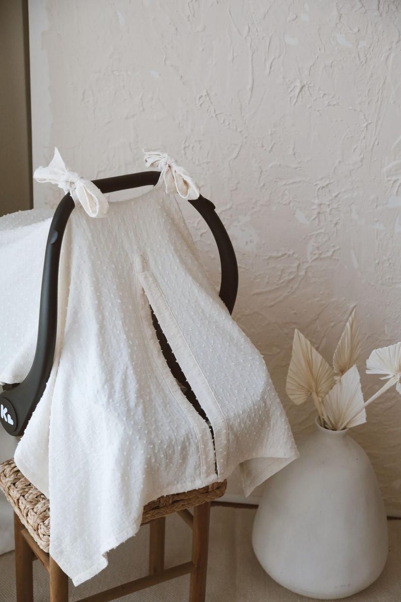 May include: A cream-colored, textured fabric car seat cover featuring a Swiss dot pattern. The cover is draped over a black car seat handle and secured with two fabric ties at the top. A vertical opening runs down the center of the front panel.