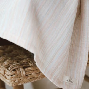 May include: A lightweight fabric blanket featuring a soft vertical stripe pattern in muted peach, light blue, and white tones with subtle metallic silver threads. A small rectangular white label is sewn on the corner with the text BABYKAR.