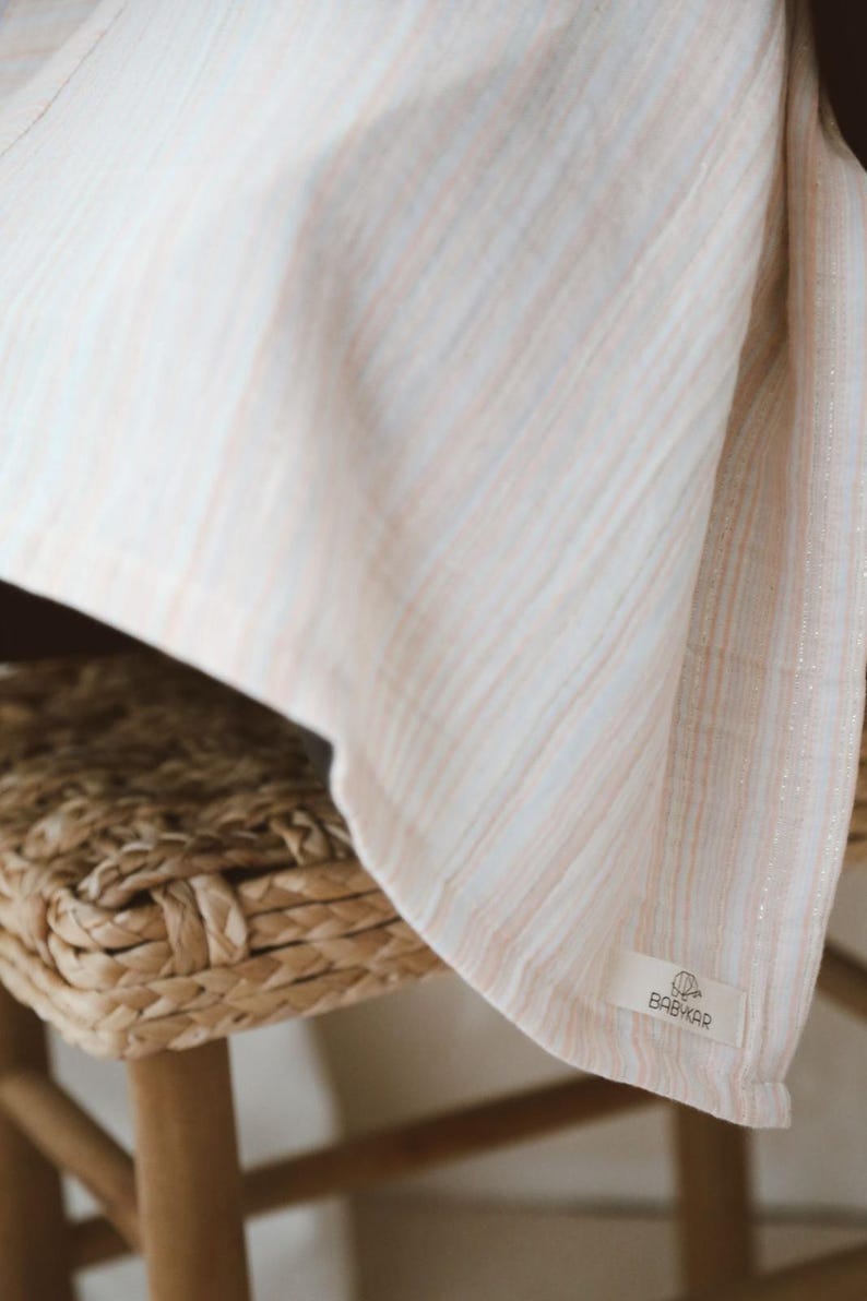 May include: A lightweight fabric blanket featuring a soft vertical stripe pattern in muted peach, light blue, and white tones with subtle metallic silver threads. A small rectangular white label is sewn on the corner with the text BABYKAR.