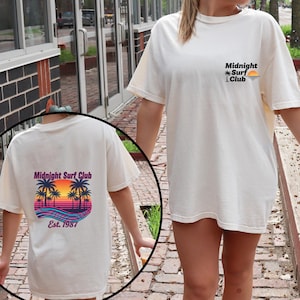 May include: Cream-colored t-shirt with the text "Midnight Surf Club" and a sunset graphic on the back. The front of the shirt has the same text and a smaller graphic. The shirt is a casual, short-sleeved style.