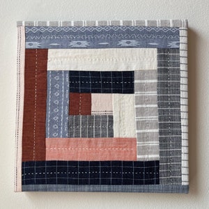 May include: A square quilt with a log cabin pattern in shades of blue, gray, white, pink, and brown. The quilt is made of different fabrics with various textures and patterns.