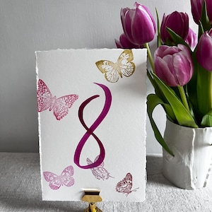May include: A rectangular white card with deckled edges featuring a large magenta number 8 in the center, surrounded by several printed butterflies in shades of magenta and gold. The card is held upright by a small gold metal stand.