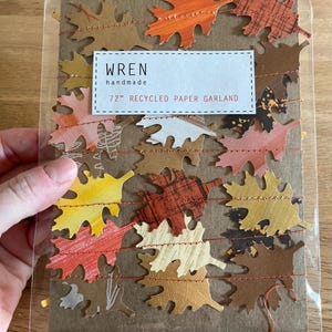 K&ouml;nnte beinhalten: Eine 183 cm lange Girlande aus recyceltem Papier mit bunten Herbstbl&auml;ttern in Orange, Gelb, Braun und Creme. Die Bl&auml;tter sind mit rotem Faden aufgereiht. Die Verpackung tr&auml;gt den Text "wren handmade".
