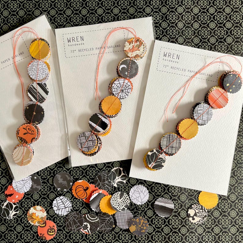 May include: Three Halloween paper garlands with orange, black, and white circular designs. Each garland is attached to a white card with the text "Wren handmade 72" Recycled Paper Garland". Loose paper circles with Halloween patterns are scattered below.