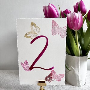 May include: A rectangular white card with deckled edges featuring a large dark pink number 2 in the center. The card is decorated with one gold butterfly and four pink butterflies in various sizes. It is held upright by a small gold metal stand.