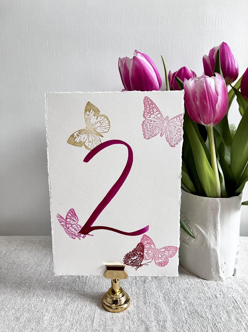 May include: A rectangular white card with deckled edges featuring a large dark pink number 2 in the center. The card is decorated with one gold butterfly and four pink butterflies in various sizes. It is held upright by a small gold metal stand.