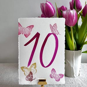 May include: A rectangular white card with deckled edges featuring the number 10 in a large magenta font. The card is decorated with three magenta butterfly illustrations and one gold butterfly illustration, held upright by a gold metal stand.
