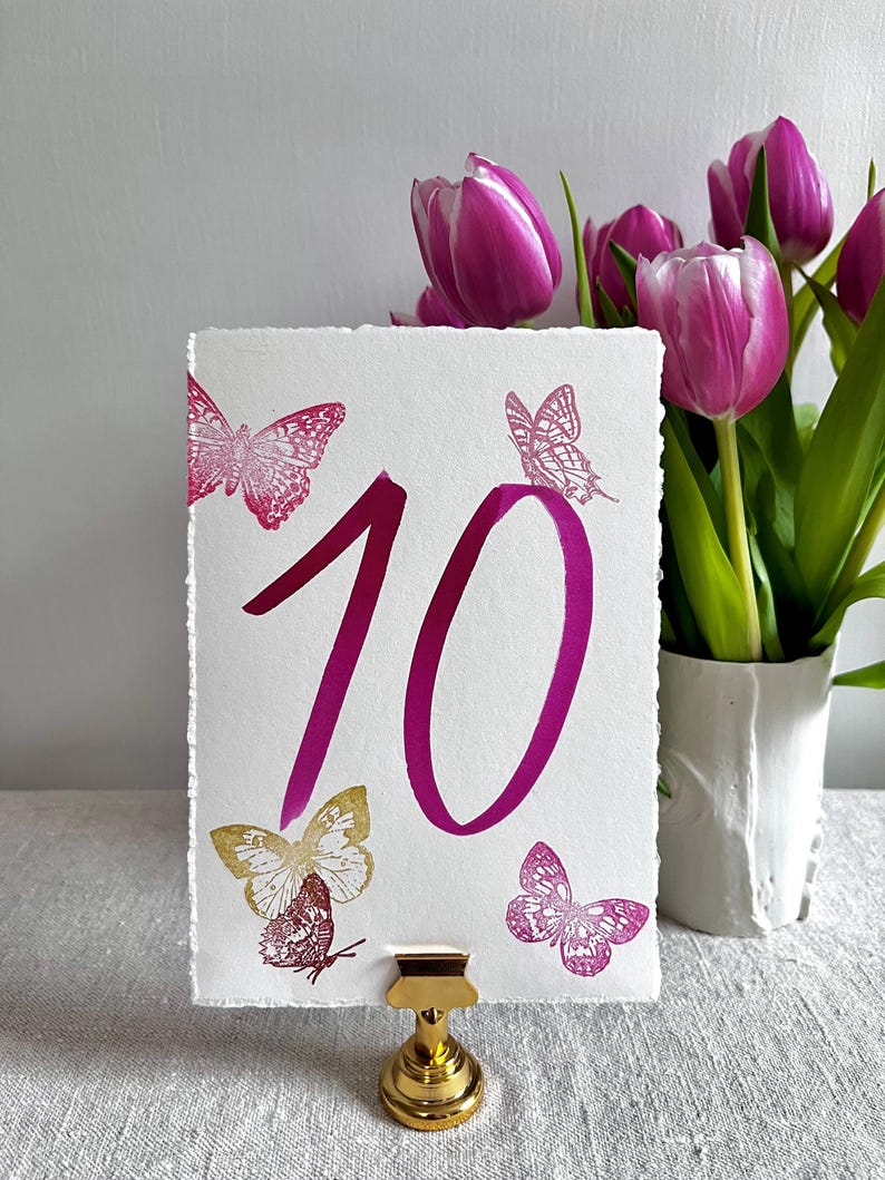 May include: A rectangular white card with deckled edges featuring the number 10 in a large magenta font. The card is decorated with three magenta butterfly illustrations and one gold butterfly illustration, held upright by a gold metal stand.